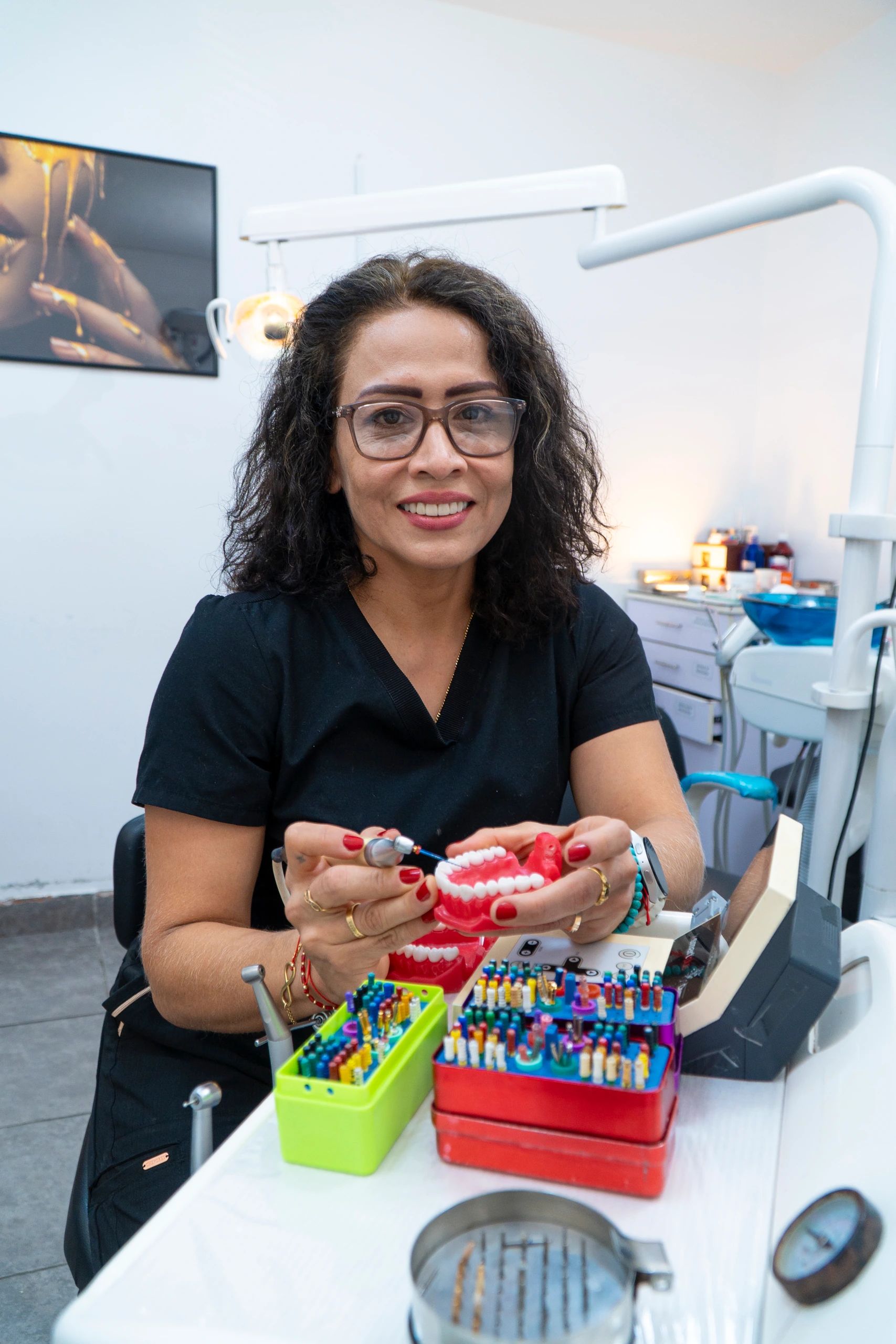 Especialista sonriente sostiene un modelo dental y una lima, frente a las cajas de instrumental de colores sobre la mesa del consultorio.