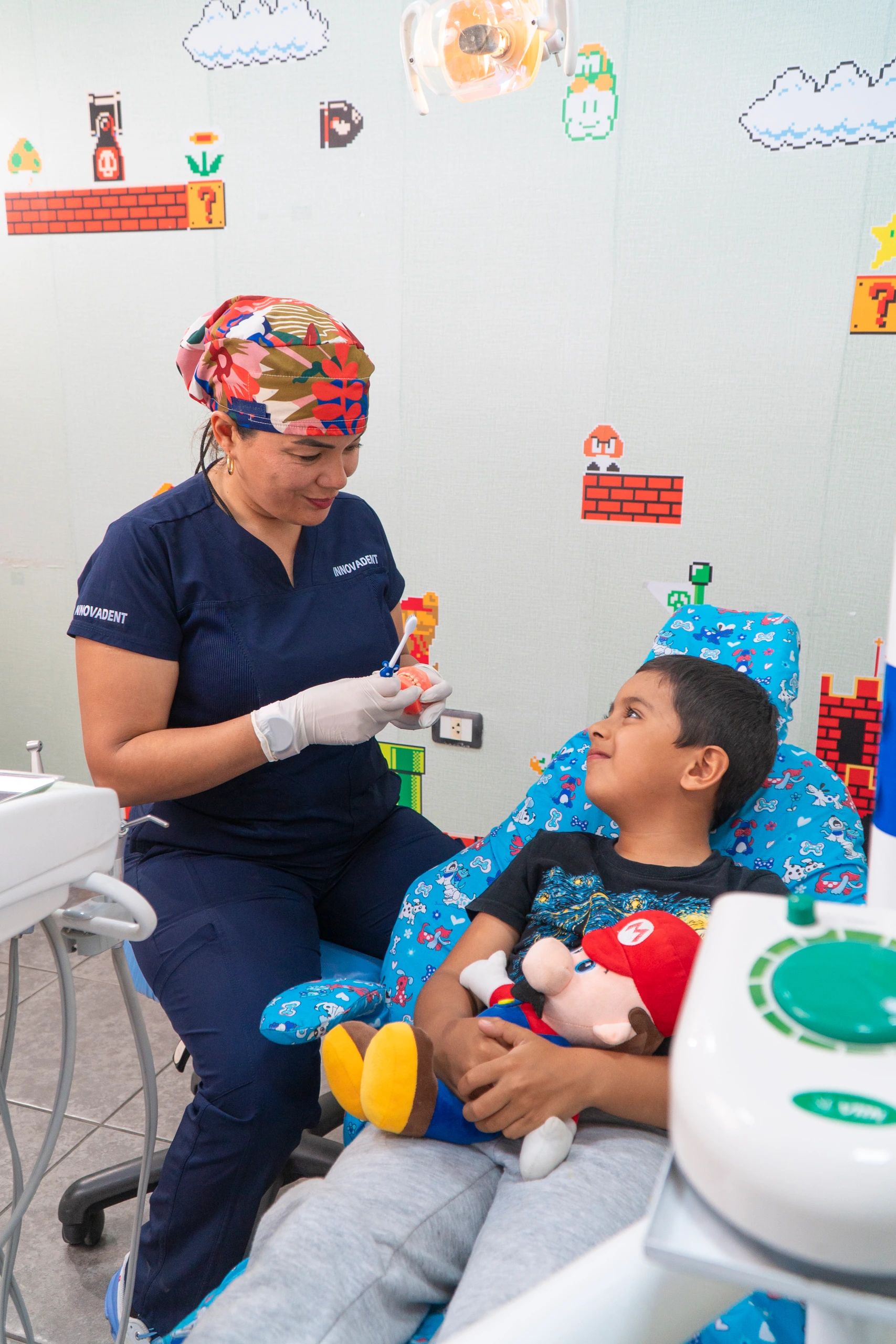 La odontopediatra conversa con un niño que abraza un peluche en el sillón infantil, mientras le muestra un cepillo y un modelo dental