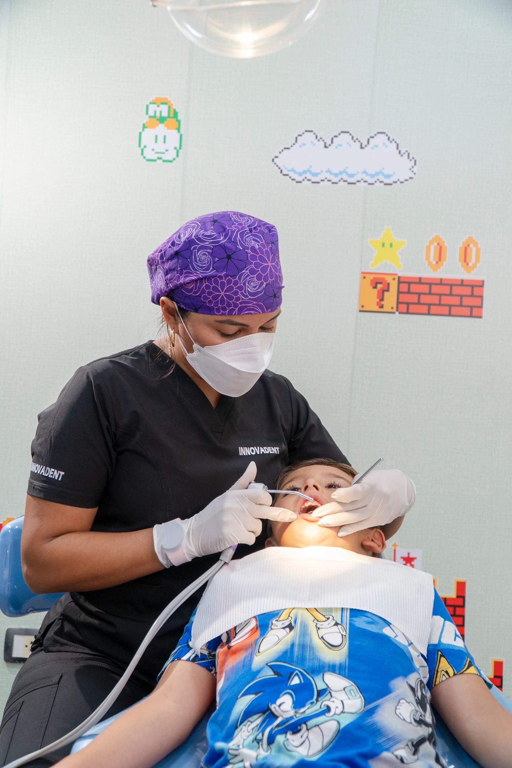 La odontopediatra revisa los dientes de un niño recostado en el sillón, frente a una pared con figuras de videojuego