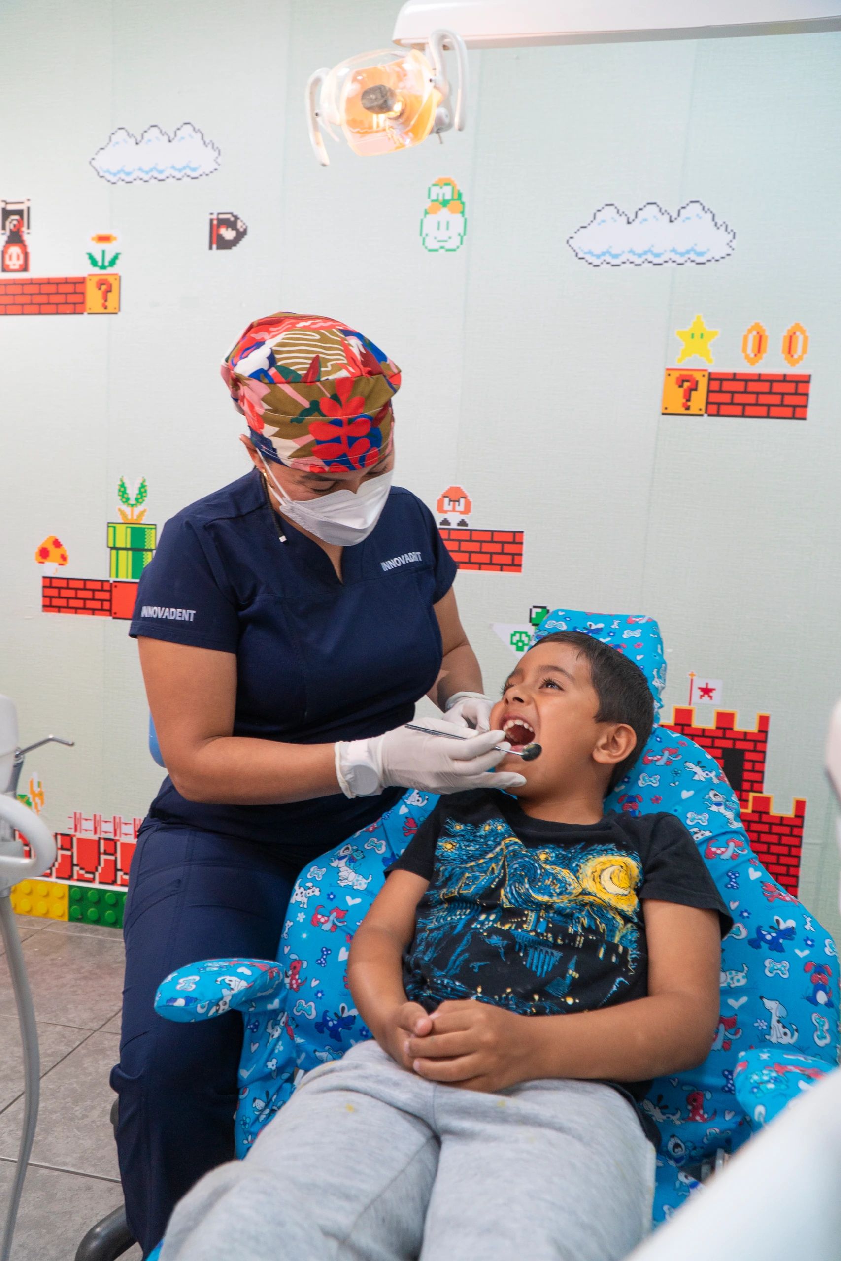 La odontopediatra revisa la boca de un niño sentado en un sillón infantil, en el consultorio decorado con figuras de videojuego