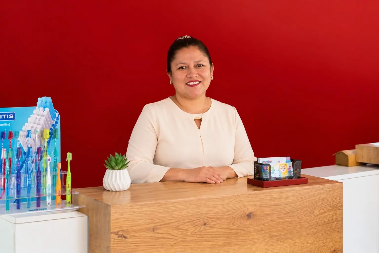La recepcionista sonríe detrás del mostrador de madera, frente a una pared roja y junto a un exhibidor de cepillos dentales